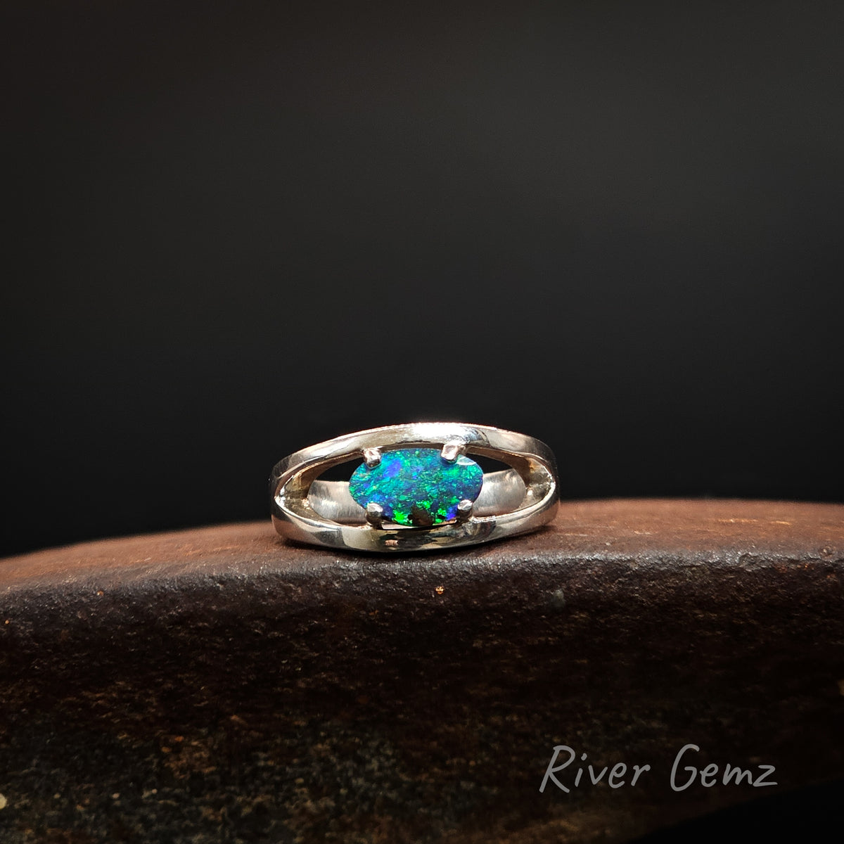 A sterling silver ring with an oval-cut, vibrant blue-green opal gemstone, displayed on a dark textured surface against a black background. The brand name 'River Gemz' is visible in the bottom right corner.