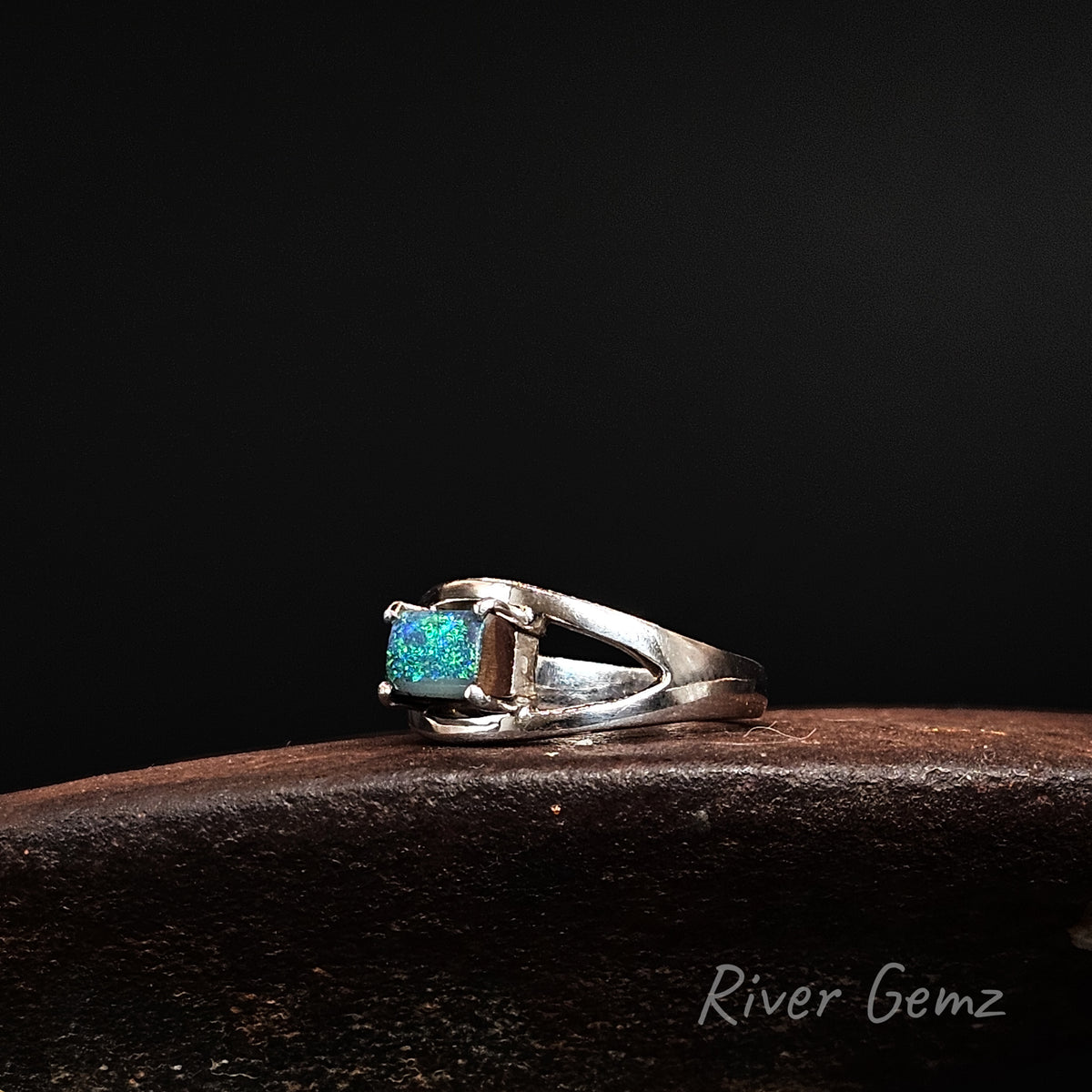 A sterling silver ring featuring a rectangular blue opal gemstone, displayed on a dark brown wooden surface against a solid black background. The brand name 'River Gemz' is visible as a watermark in the bottom right corner.