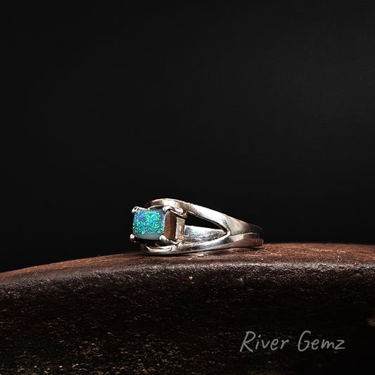 A sterling silver ring featuring a rectangular blue opal gemstone, displayed on a dark brown wooden surface against a solid black background. The brand name 'River Gemz' is visible as a watermark in the bottom right corner.