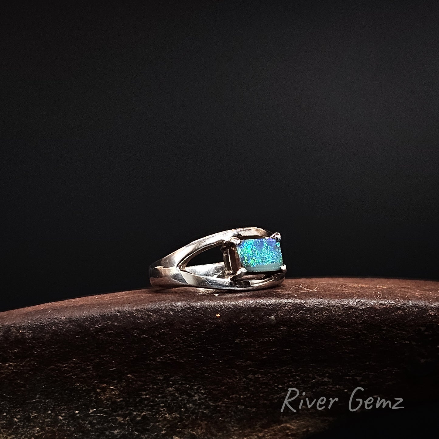 A sterling silver ring with a rectangular blue opal gemstone, resting on a dark brown textured surface against a solid black background. The brand name 'River Gemz' is visible in the bottom right corner.