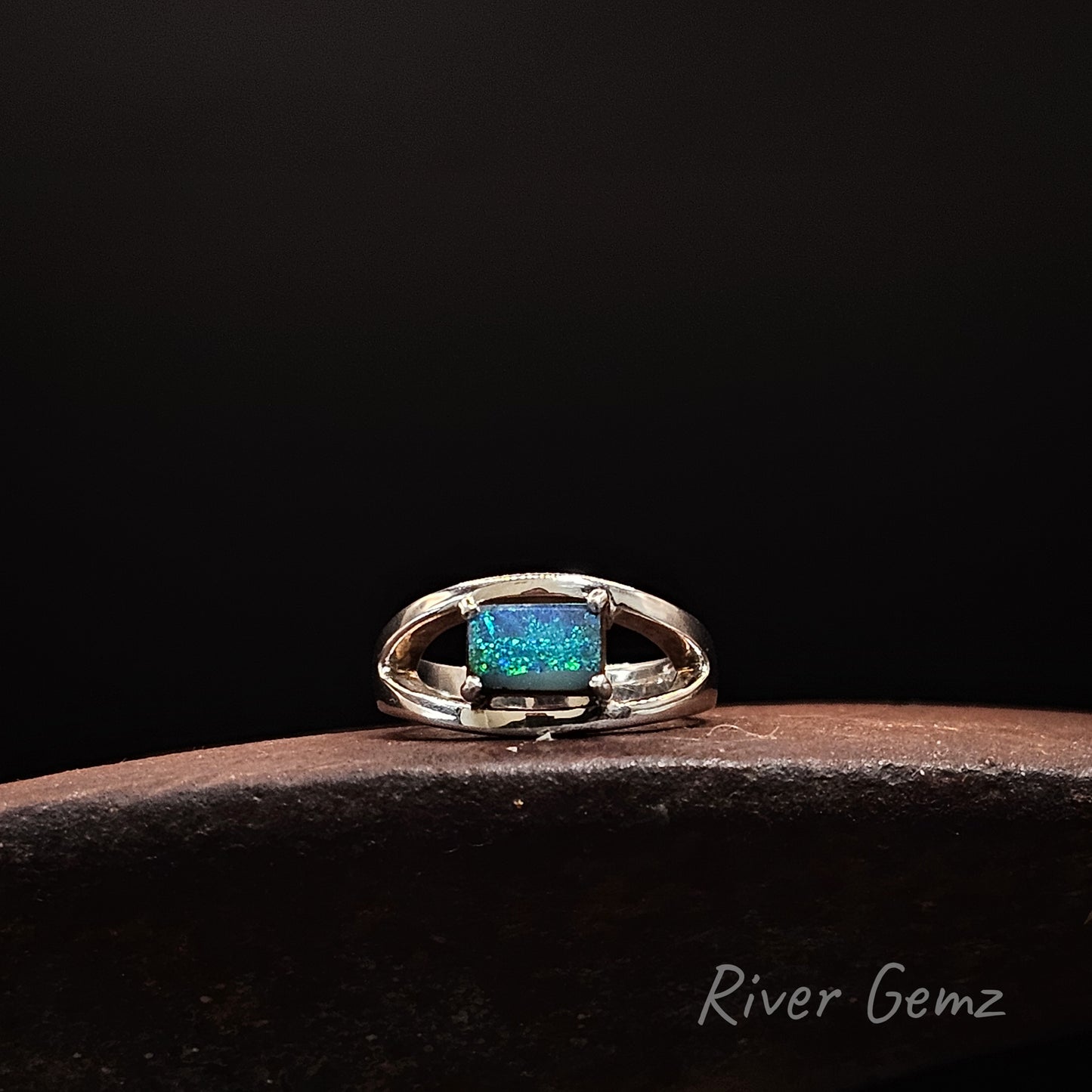 A silver ring featuring a rectangular blue-green boulder opal, resting on a dark brown wooden surface against a solid black background. The brand name 'River Gemz' is visible in the bottom right corner.