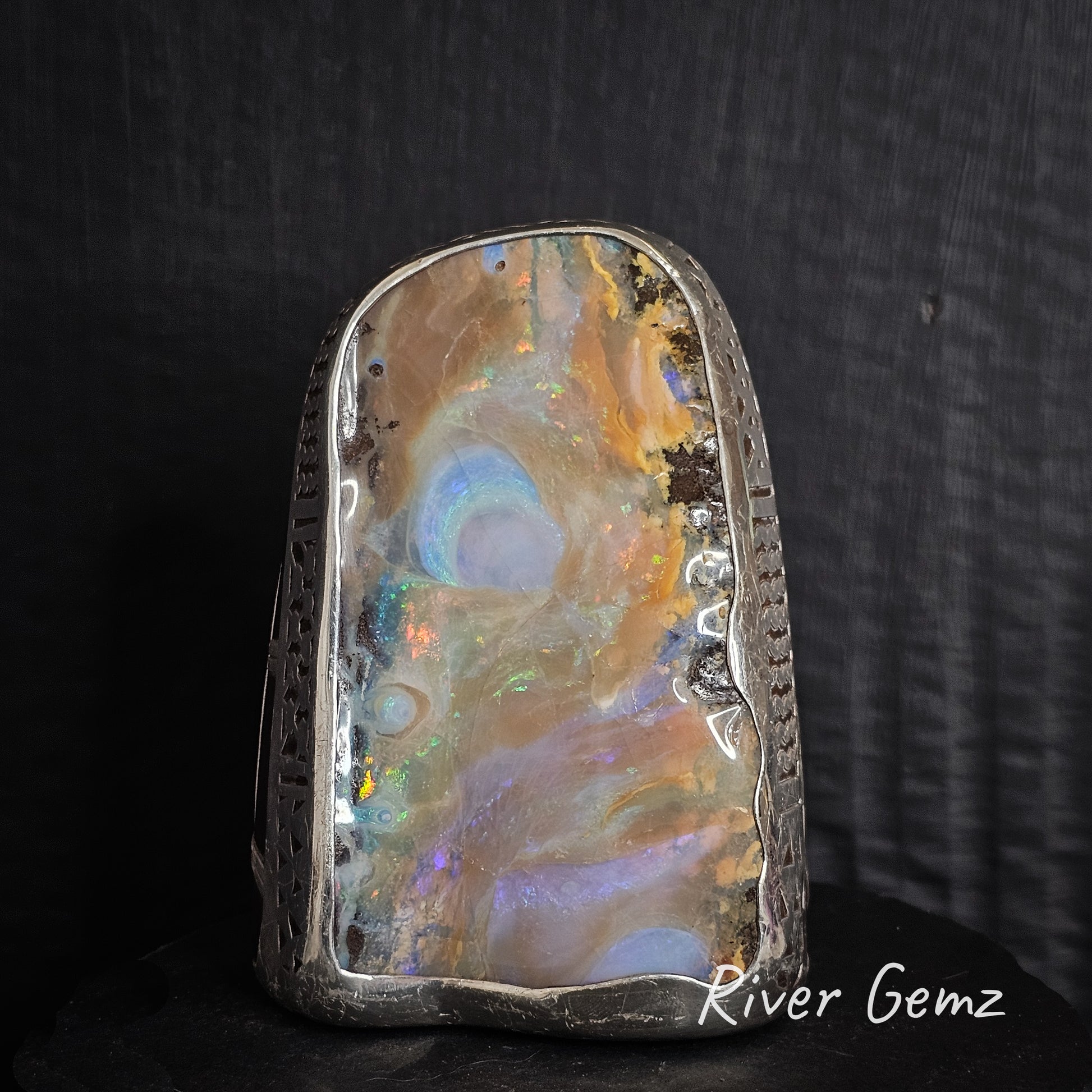 Opal stone with colorful swirling patterns on a dark background, branded 'River Gemz'.