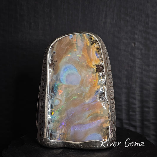 Opal stone with colorful swirling patterns on a dark background, branded 'River Gemz'.