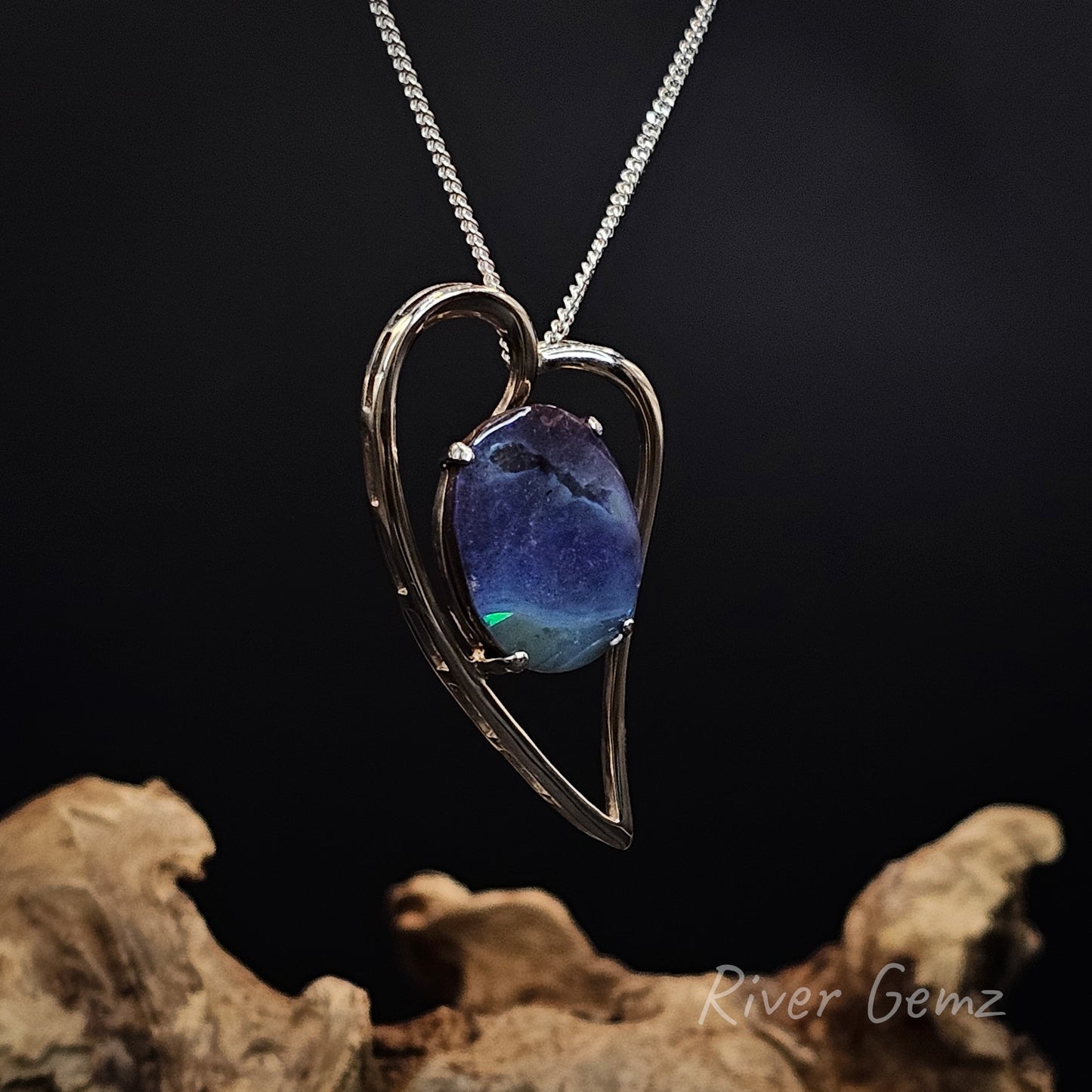 Heart-shaped pendant with a blue stone on a chain, set against a dark background with rocks.