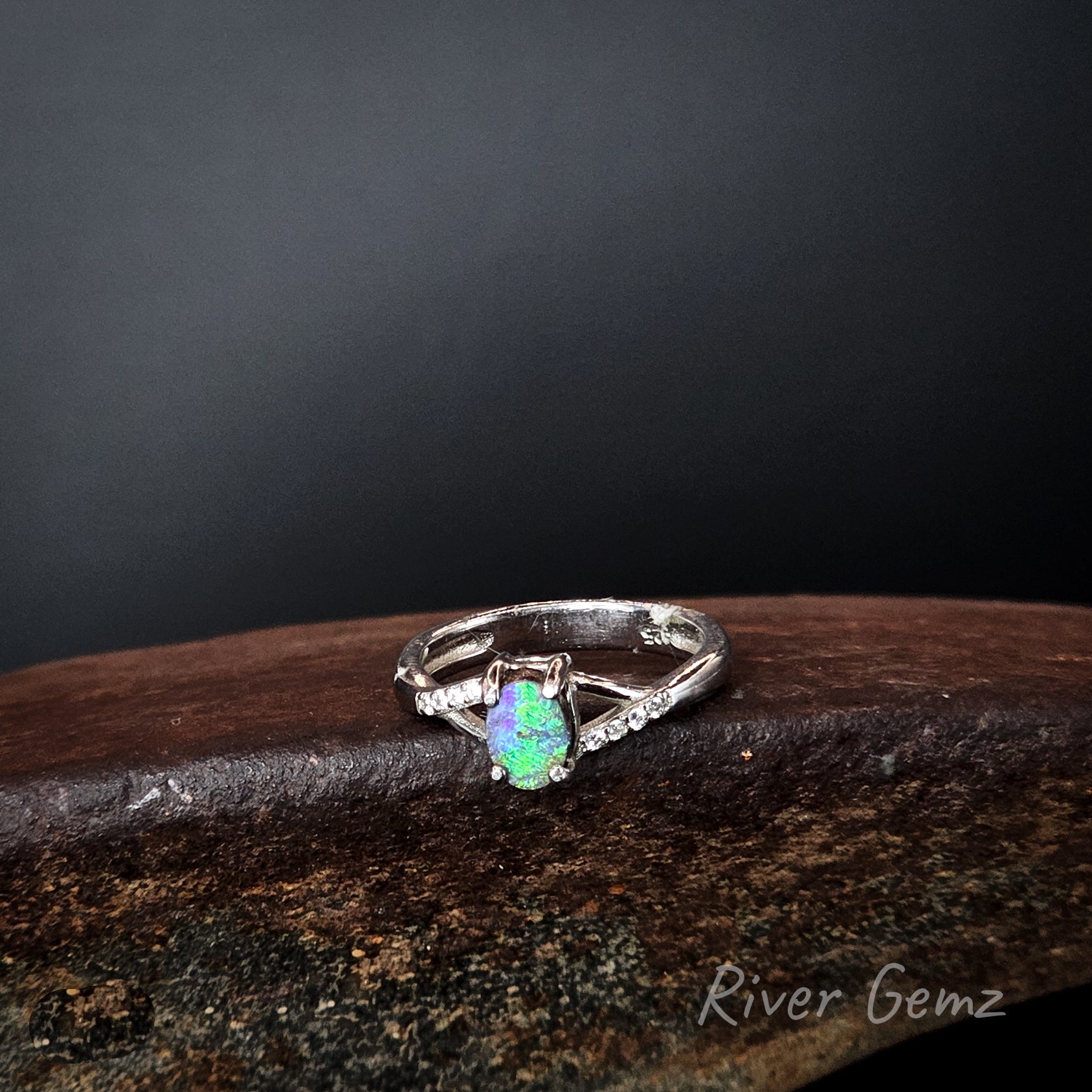 Green opal set in silver ring on a rusty coloured surface with 'River Gemz' branding.