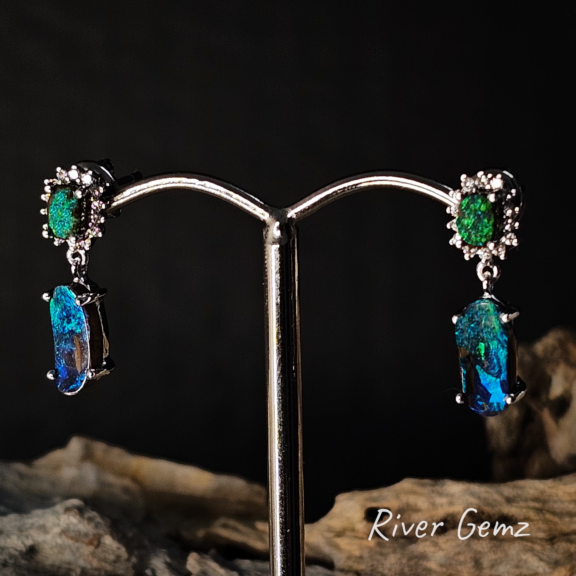 Pair of earrings with green and blue boulder opals and diamonds on a stand with a dark background.