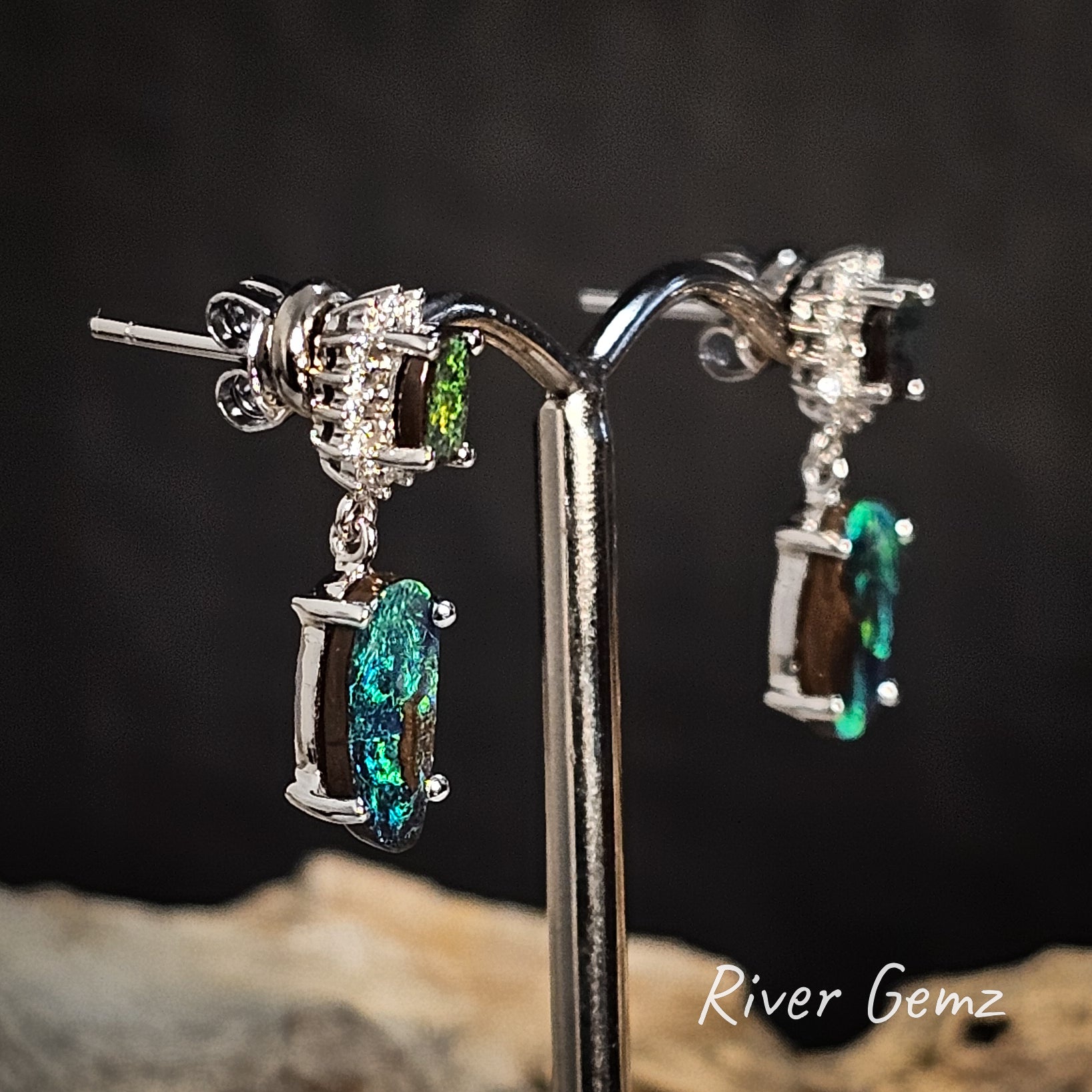 Pair of white gold earrings with two green gemstones and diamonds on a stand against a dark background, branded 'River Gemz'.