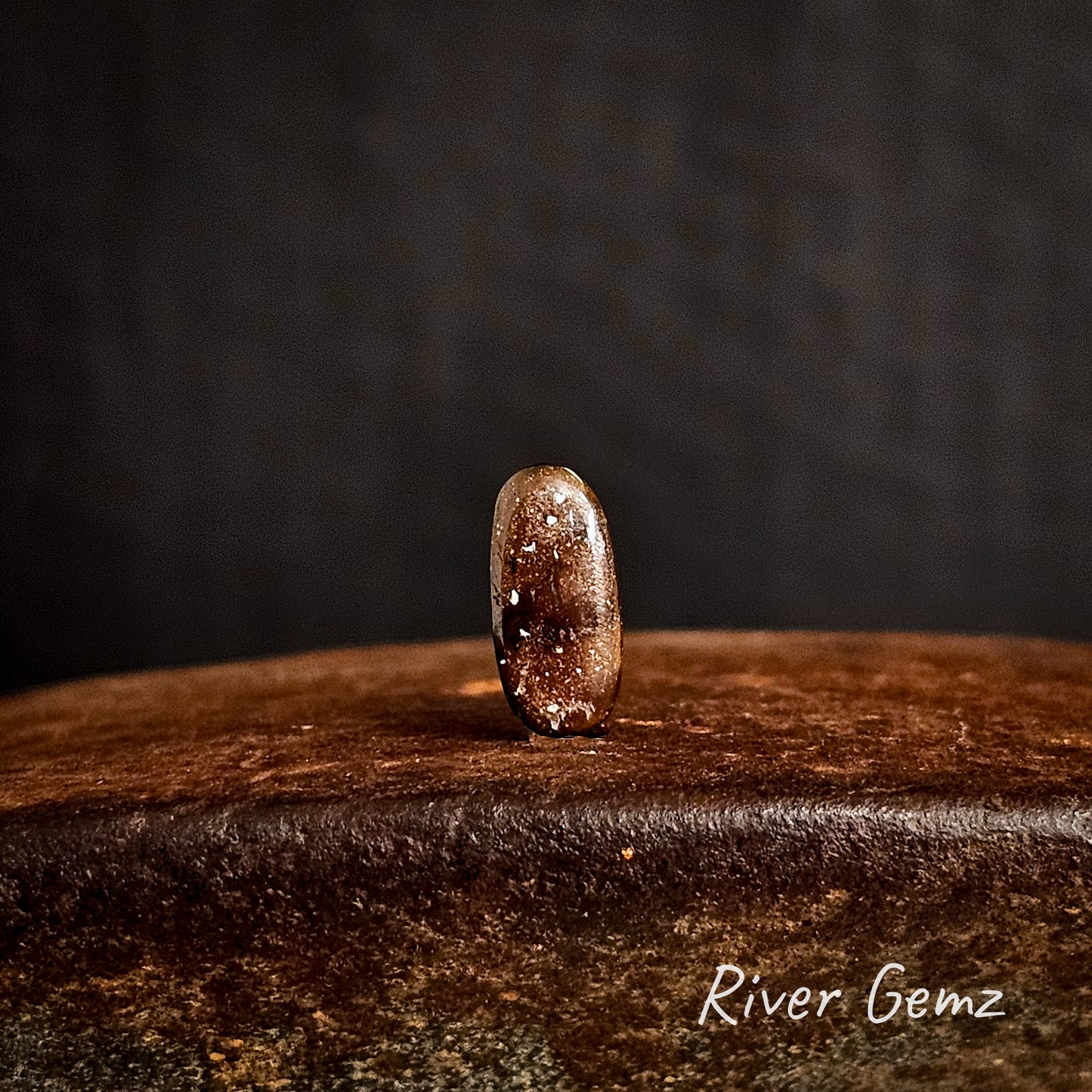 Back view of the opal shows the ironstone matrix of the boulder opal  on a cast iron surface with a dark background, featuring the brand 'River Gemz'.