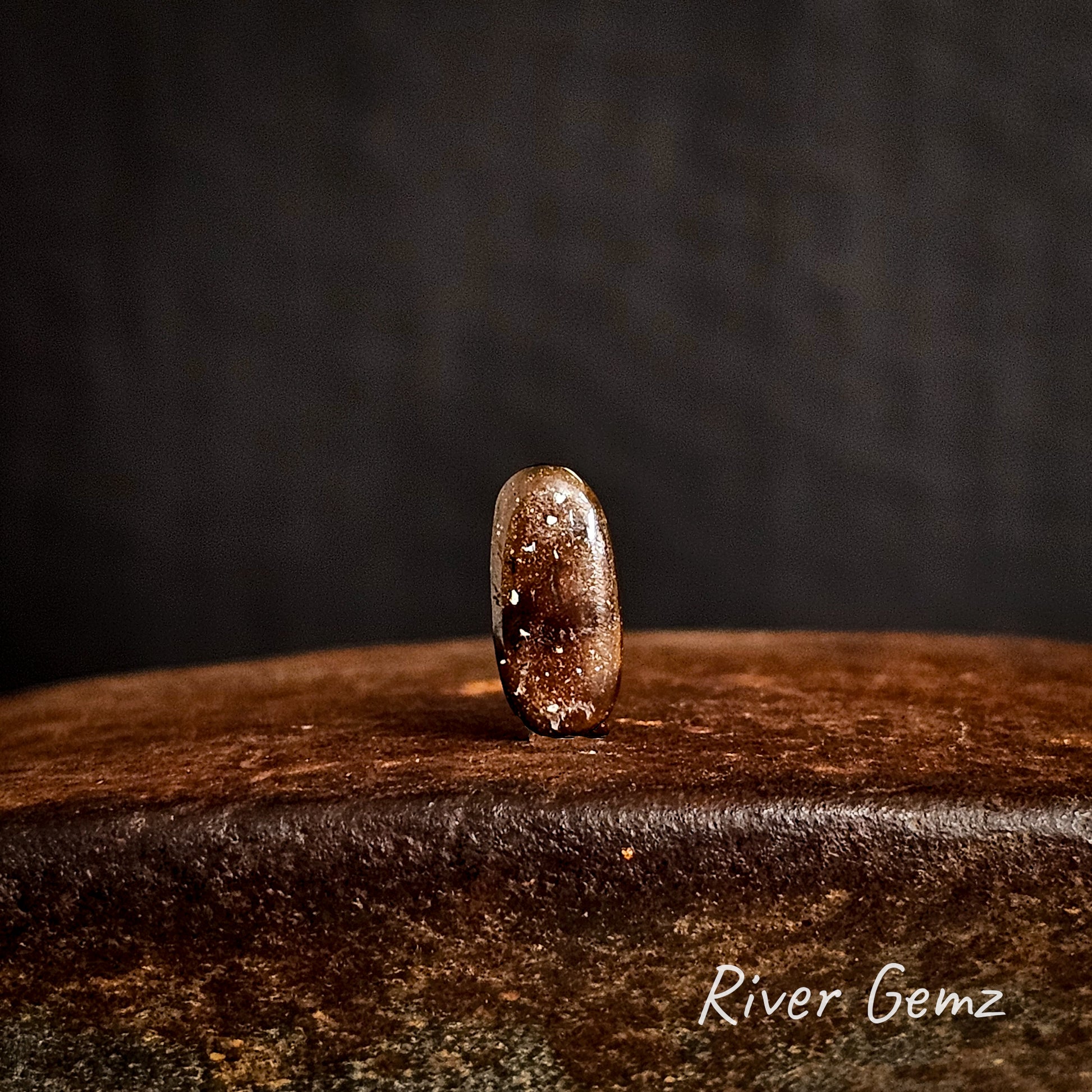 Back view of the opal shows the ironstone matrix of the boulder opal  on a cast iron surface with a dark background, featuring the brand 'River Gemz'.