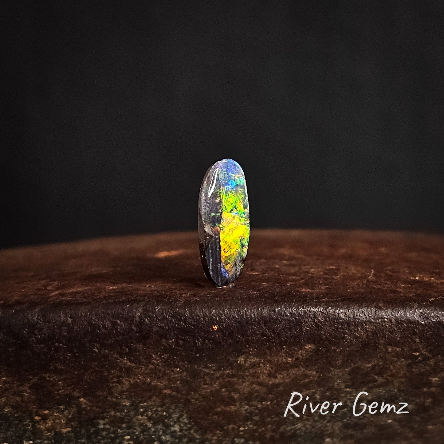 Multi-coloured oval shaped boulder opal on a dark background with 'River Gemz' branding