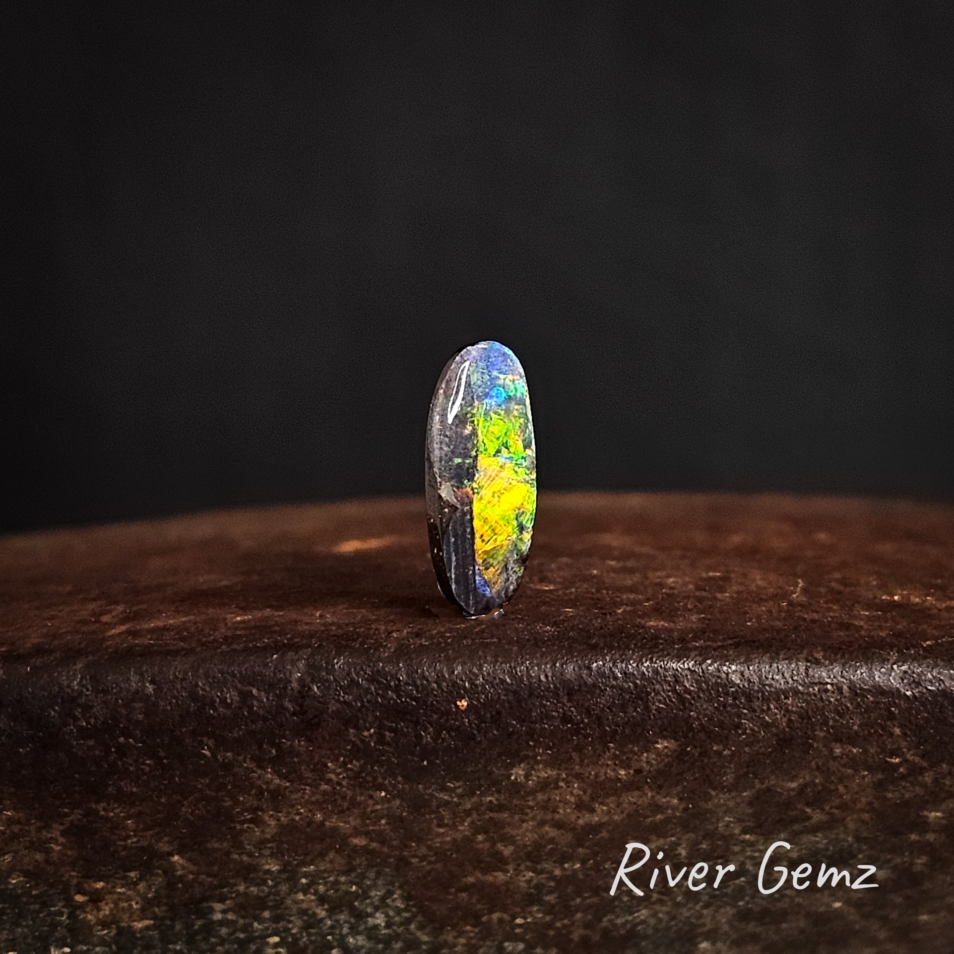 Multi-coloured oval shaped boulder opal on a dark background with 'River Gemz' branding