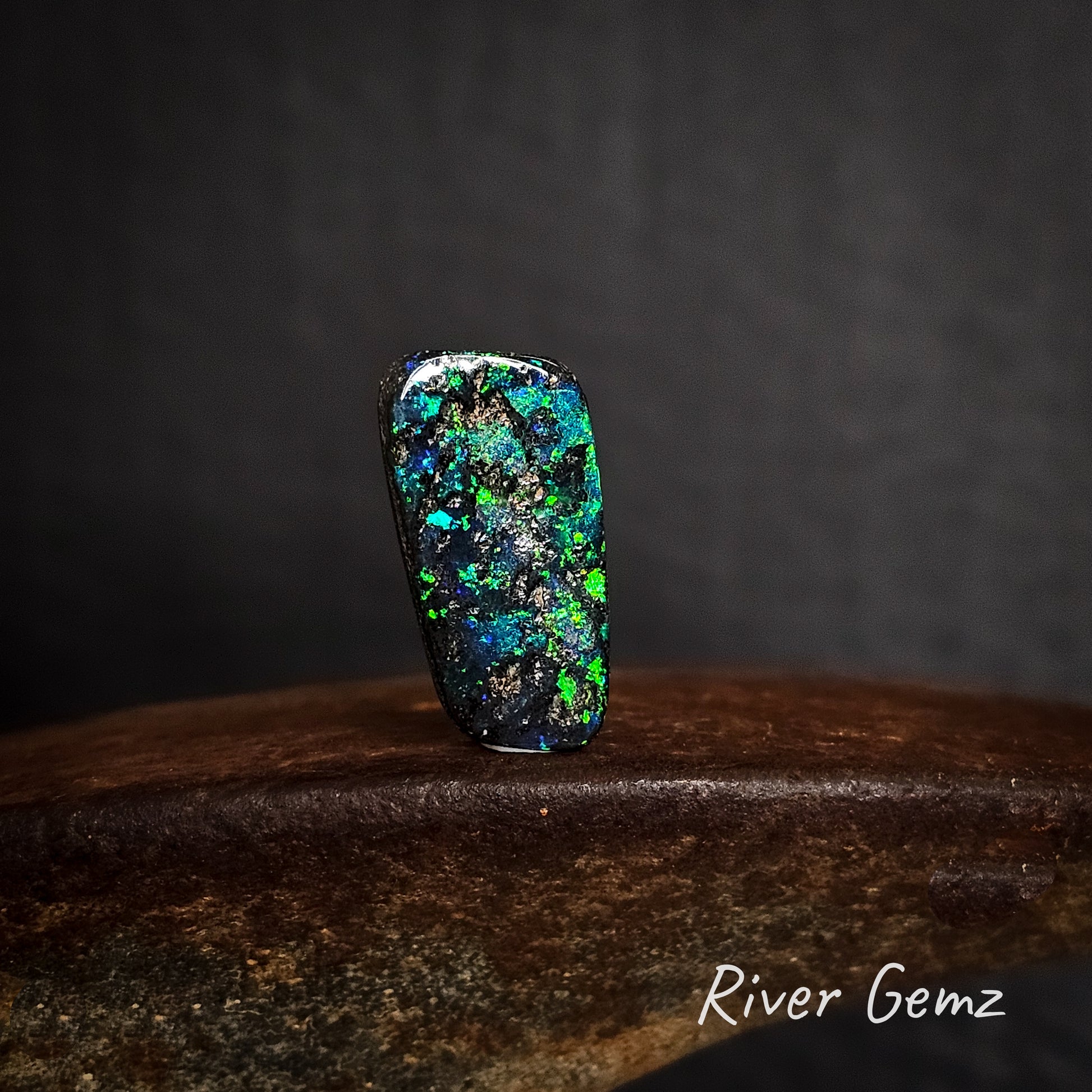 Multicolored free-form cut opal stone on a dark background with 'River Gemz' branding.