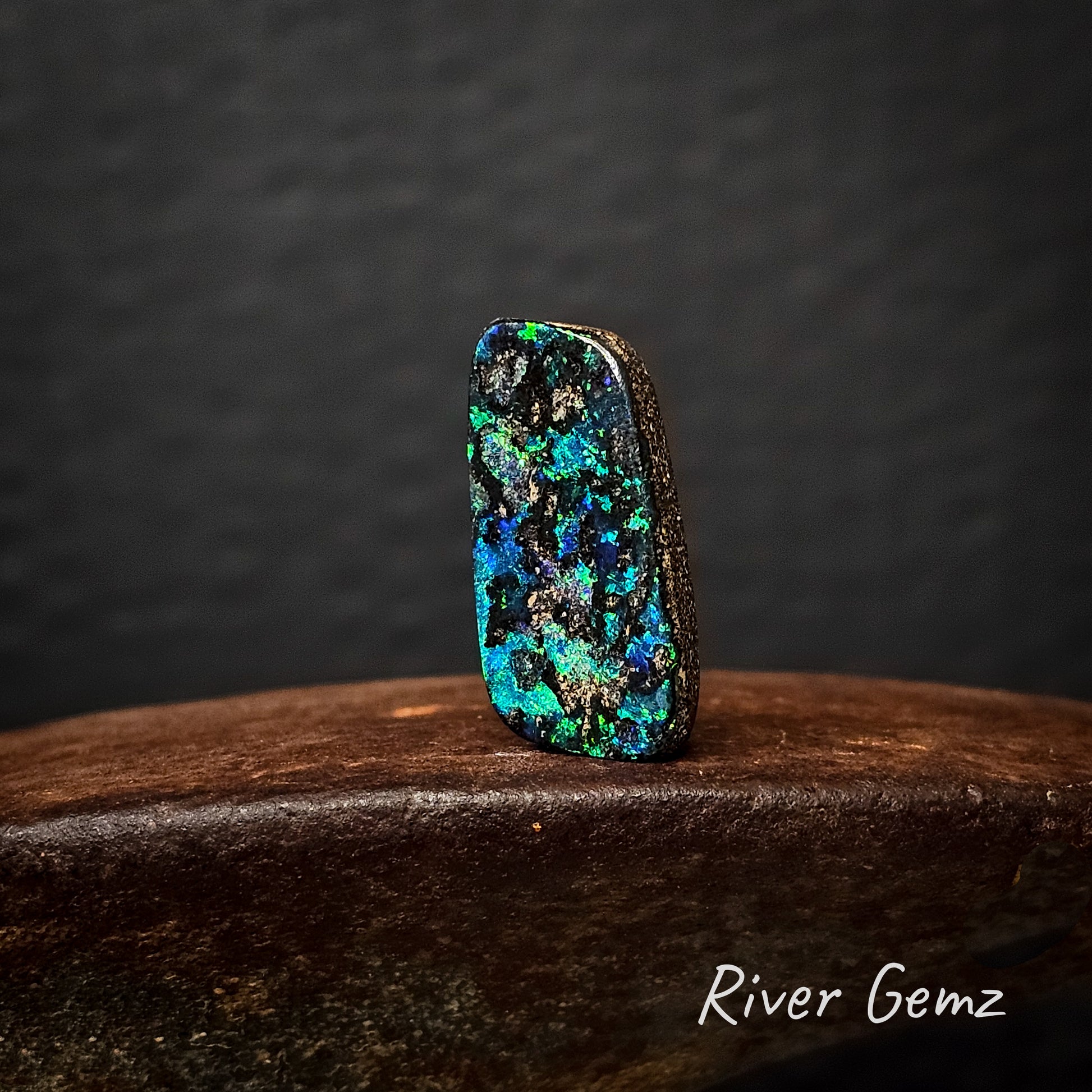 Multicolored boulder opal on a cast iron surface with a dark background, featuring the brand 'River Gemz'.
