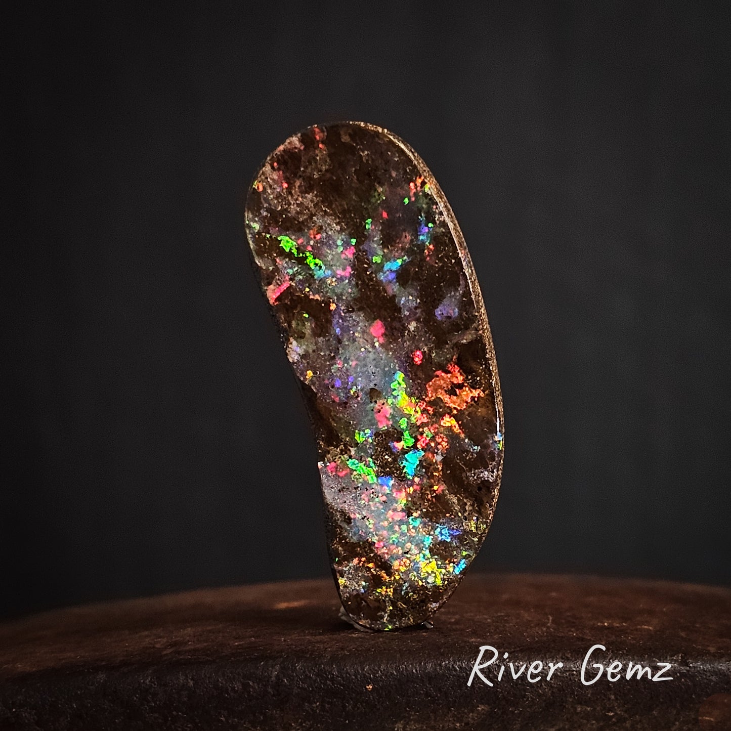 Peanut shaped colourful boulder opal standing upright against a dark background.