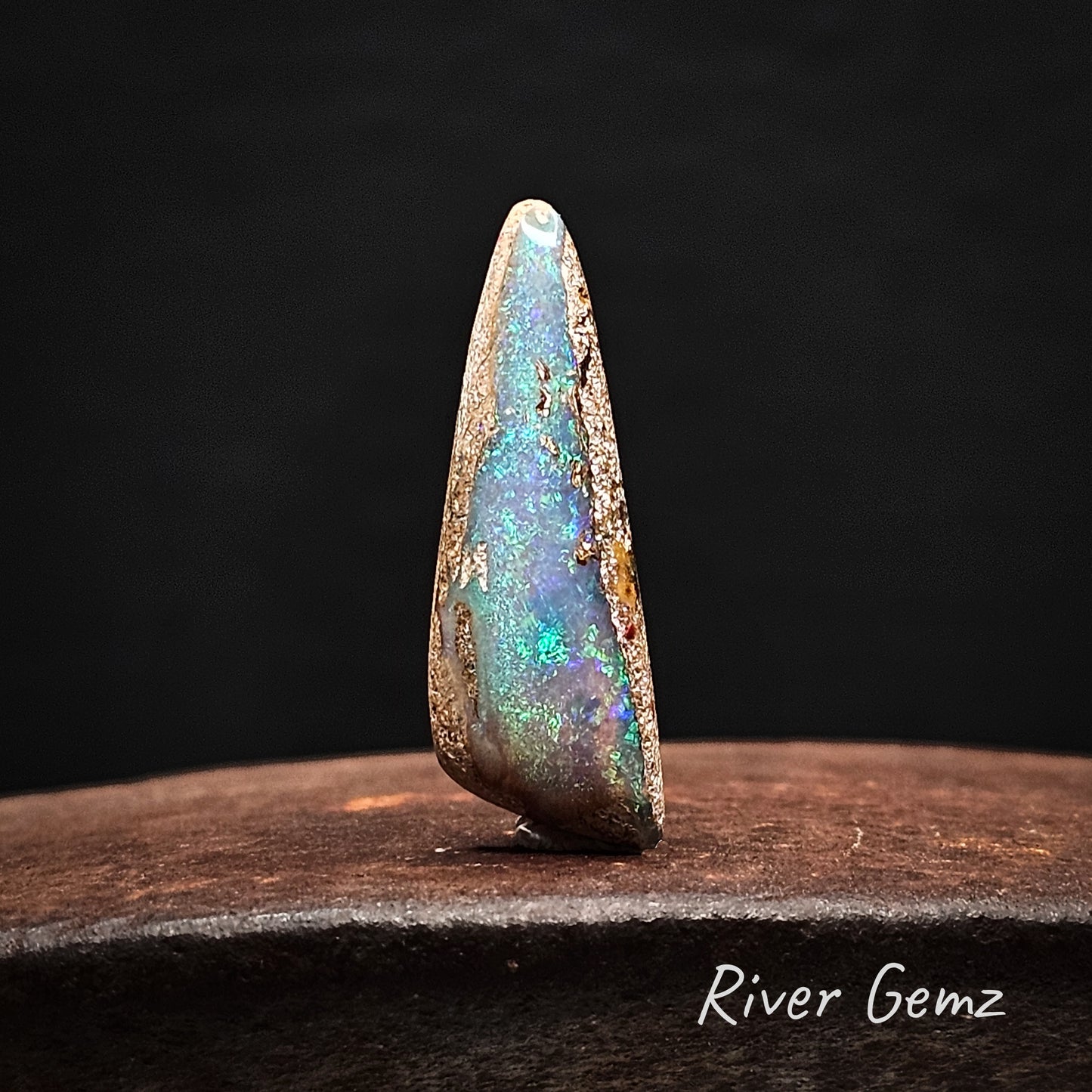 Blue and green pipe opal standing upright on an cast iron shoe anvil with 'River Gemz' branding.
