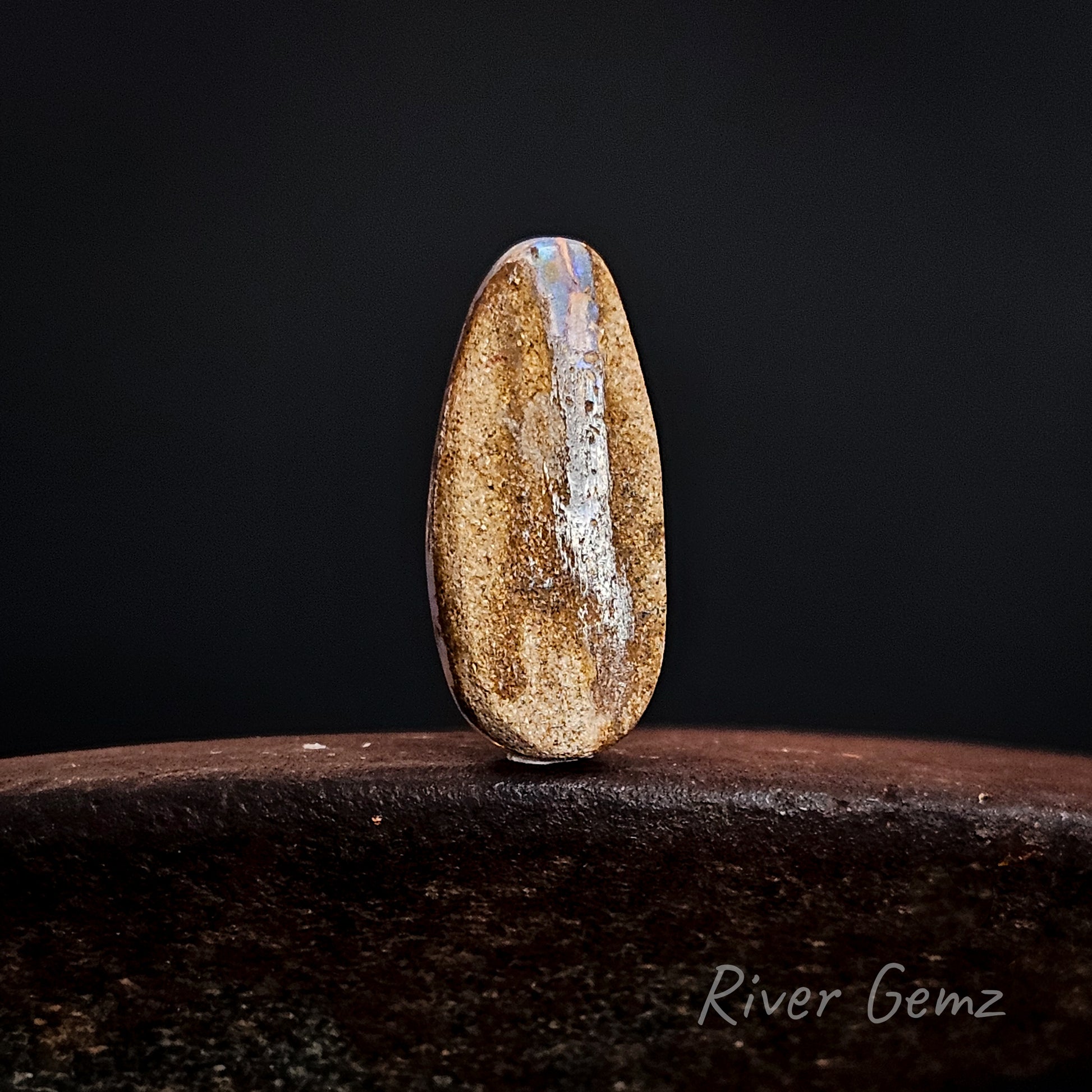 Rear view of the stone showing the light brown base tone of the pipe opal on a dark background, labeled 'River Gemz'.