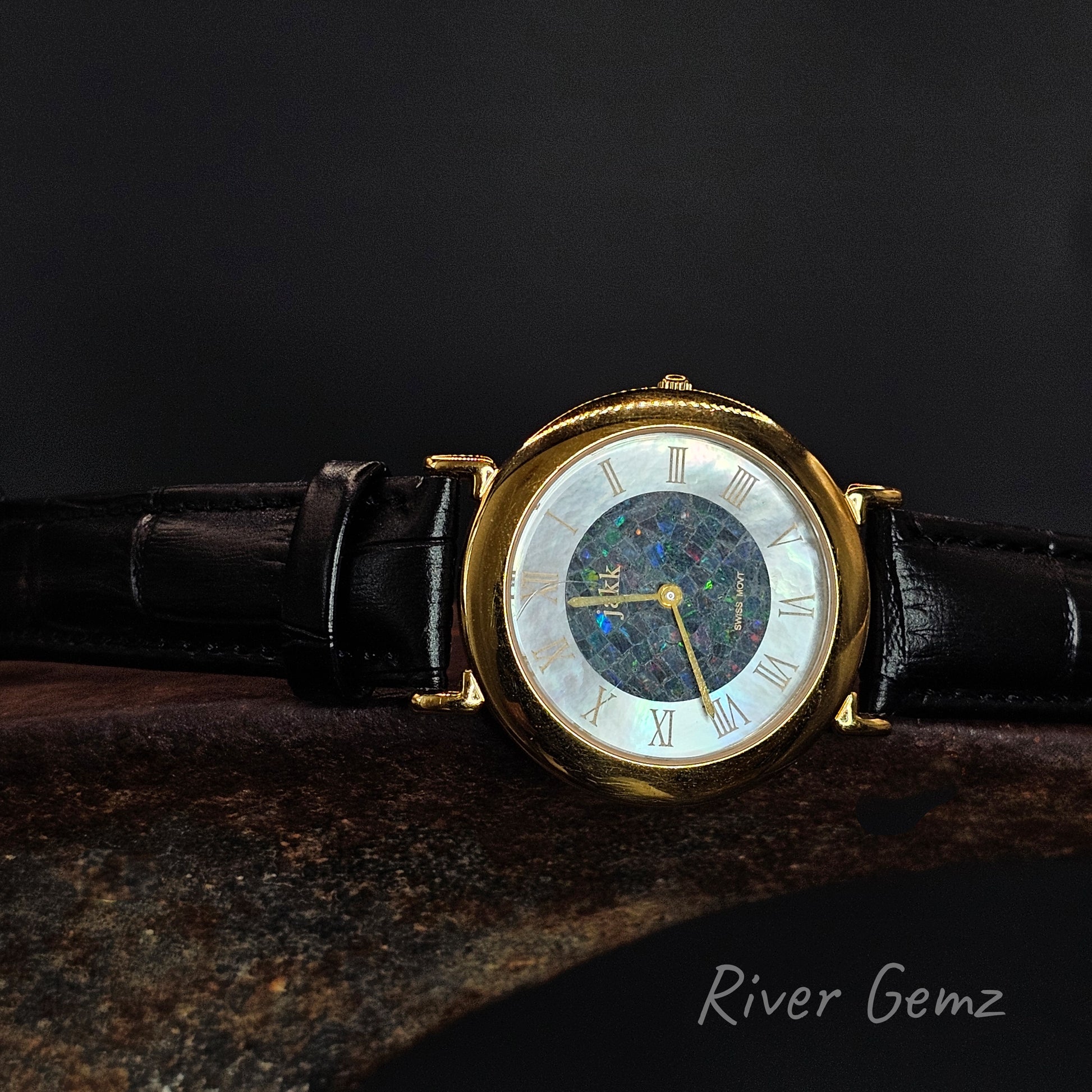 Gold wristwatch with black leather strap on a dark background, featuring the brand 'River Gemz'.