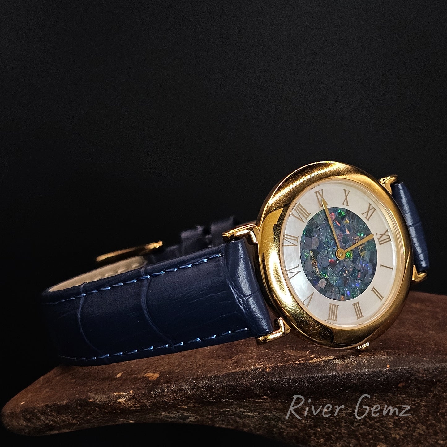 Gold wristwatch with blue leather strap on a dark background, featuring the brand 'River Gemz'.