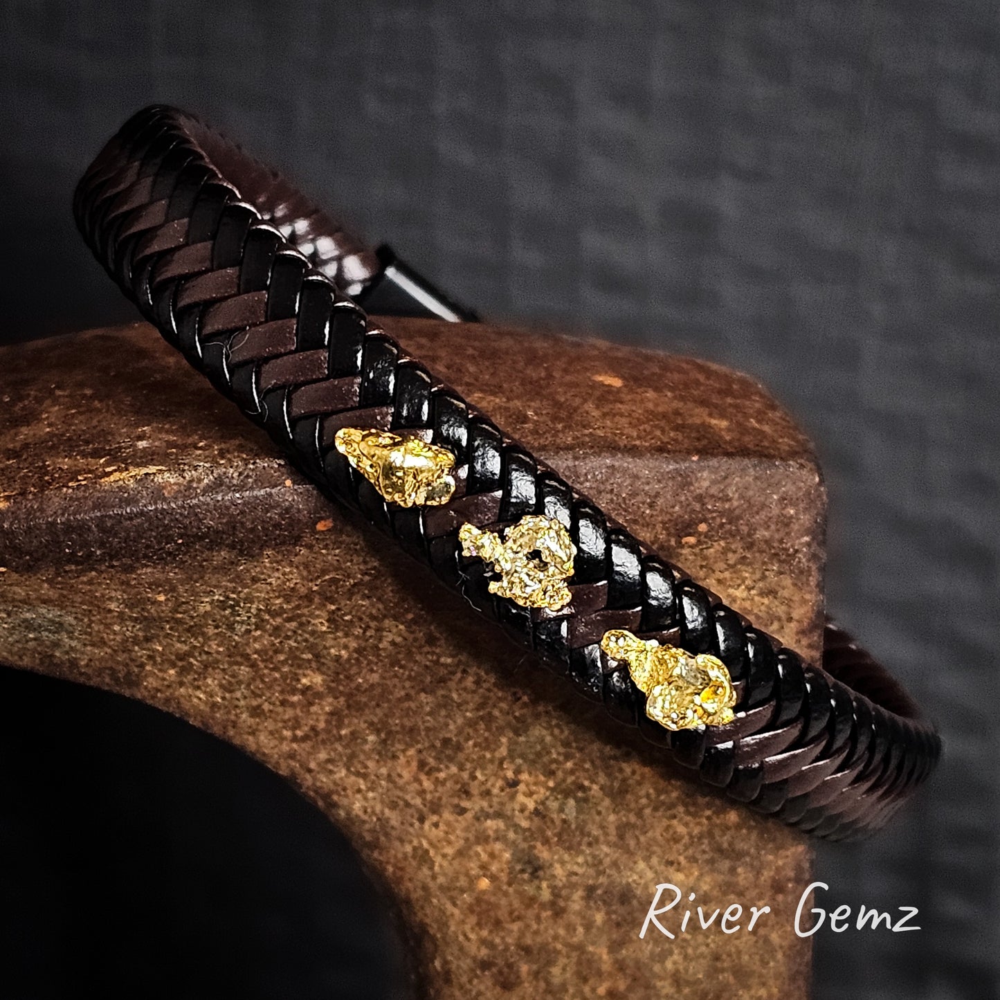 Black and brown braided leather bracelet with gold nuggets on a cast iron shoe anvil, featuring the brand 'River Gemz'.