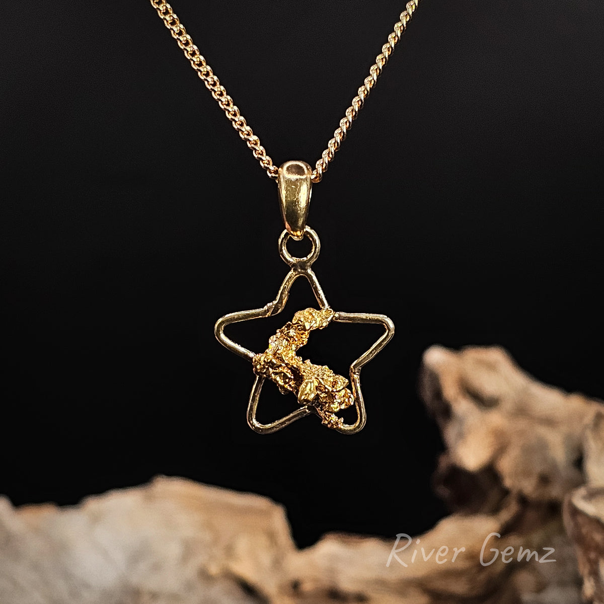 A gold star-shaped pendant necklace featuring a textured gold nugget in the center, displayed on a dark background with pieces of driftwood at the bottom. The brand name 'River Gemz' is visible in the bottom right corner.