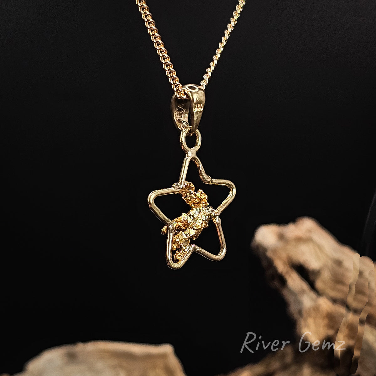 Rear view of gold nugget in a 5-pointed star shaped necklace with large bail against a black background and piece of driftwood at the base.