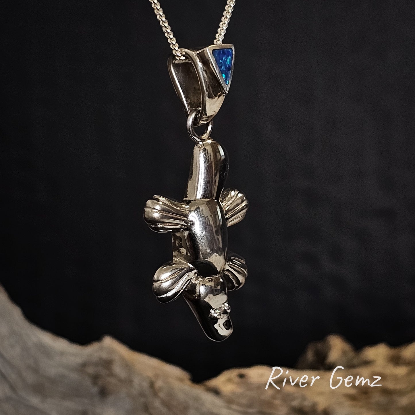 Silver platypus pendant with blue stone on a chain against a dark background, branded 'River Gemz'.