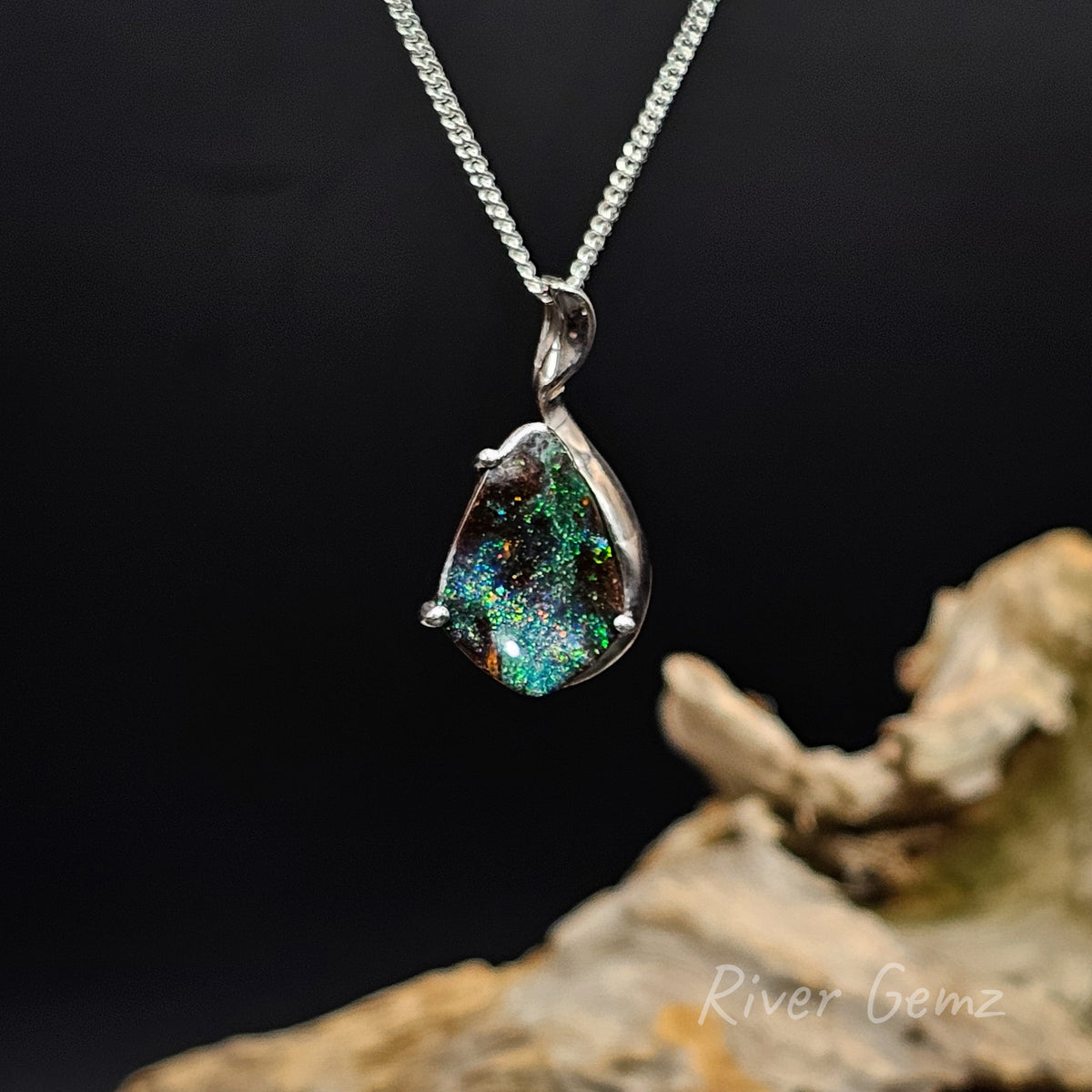 Galaxy like colours exhibited in boulder opal silver pendant against a black background and drift wood at base with 'River Gemz' water mark.