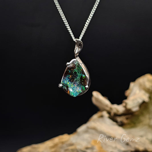 Galaxy like colours exhibited in boulder opal silver pendant against a black background and drift wood at base with 'River Gemz' water mark.