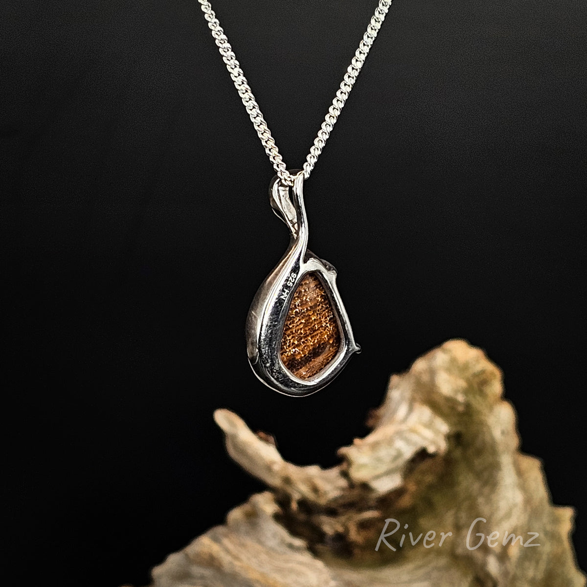 Rear view of a silver teardrop pendant necklace showing the brown base tone of the boulder opal displayed on a dark background above a piece of driftwood. The brand name 'River Gemz' is visible in the bottom right corner.