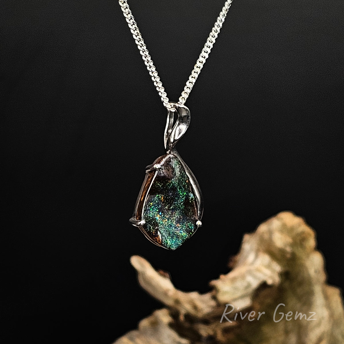 Blue and green dark opal set in sterling silver necklace against a dark background with light brown drift wood at the base showing 'River Gemz' water mark.