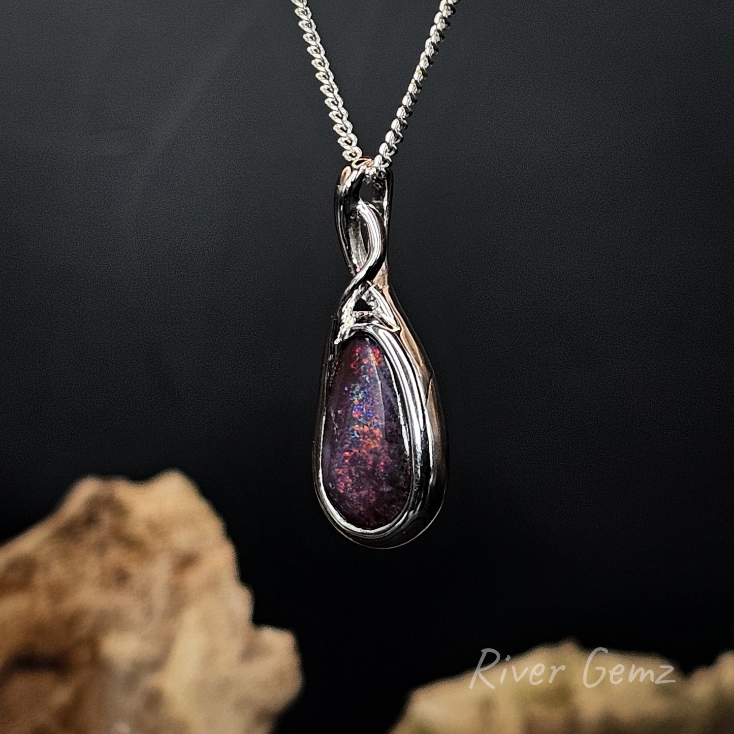 A silver teardrop pendant necklace with a swirling dual-band design, featuring a dark purple and red iridescent opal inlay, hanging against a dark background with blurred rocks at the bottom.