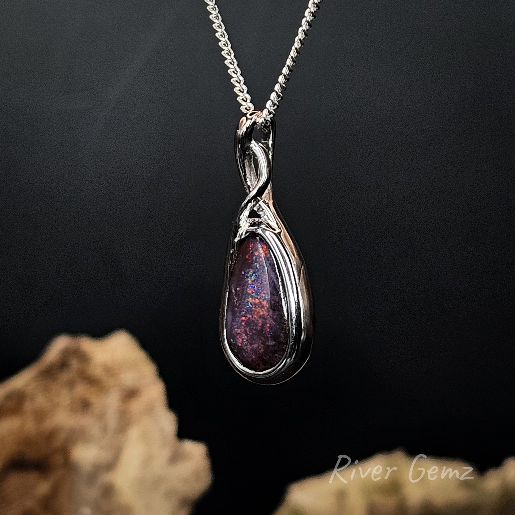 A silver teardrop pendant necklace with a swirling dual-band design, featuring a dark purple and red iridescent opal inlay, hanging against a dark background with blurred rocks at the bottom.