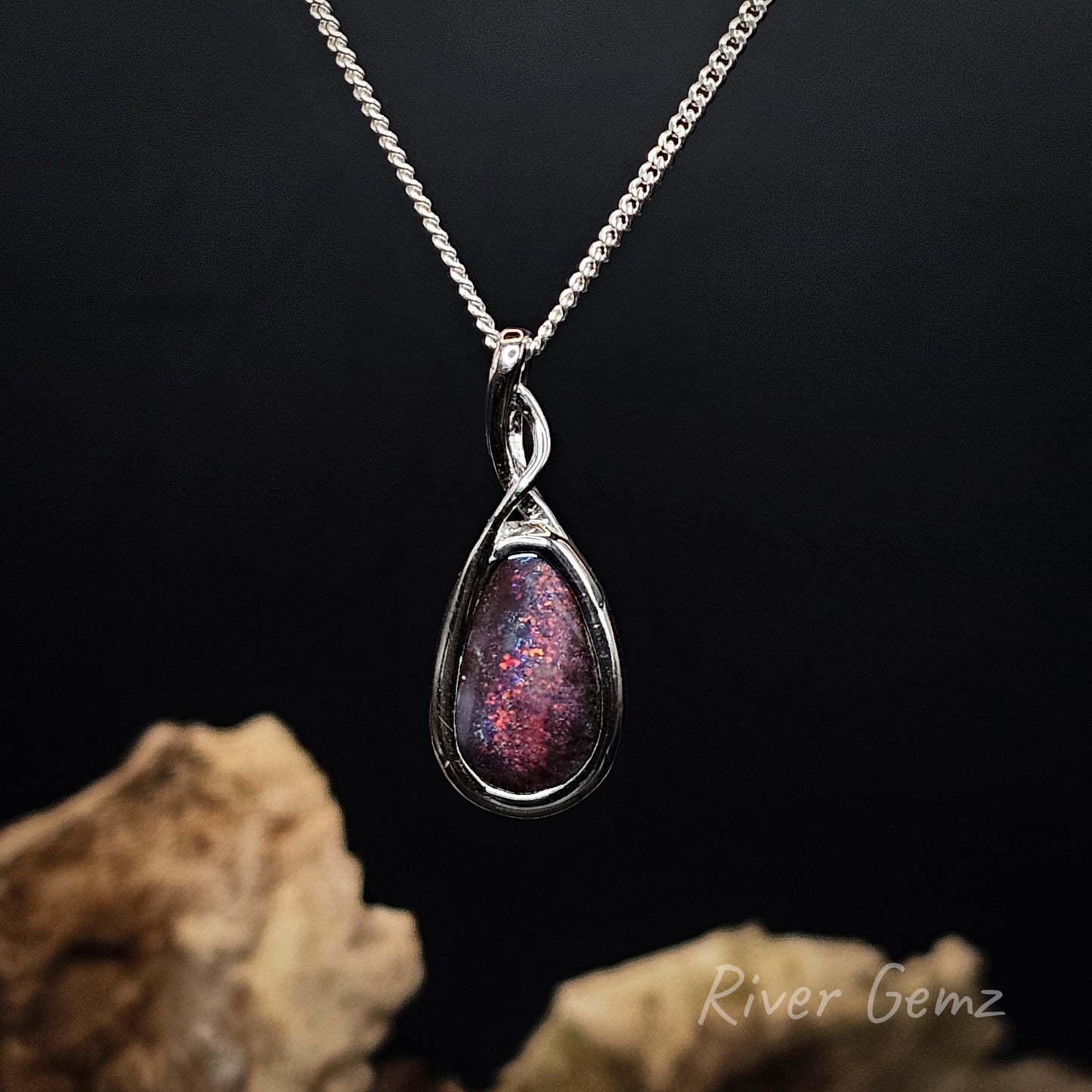 A silver teardrop pendant necklace featuring a sparkling iridescent purple and blue gemstone, suspended from a silver box chain against a dark background with decorative rocks at the bottom.