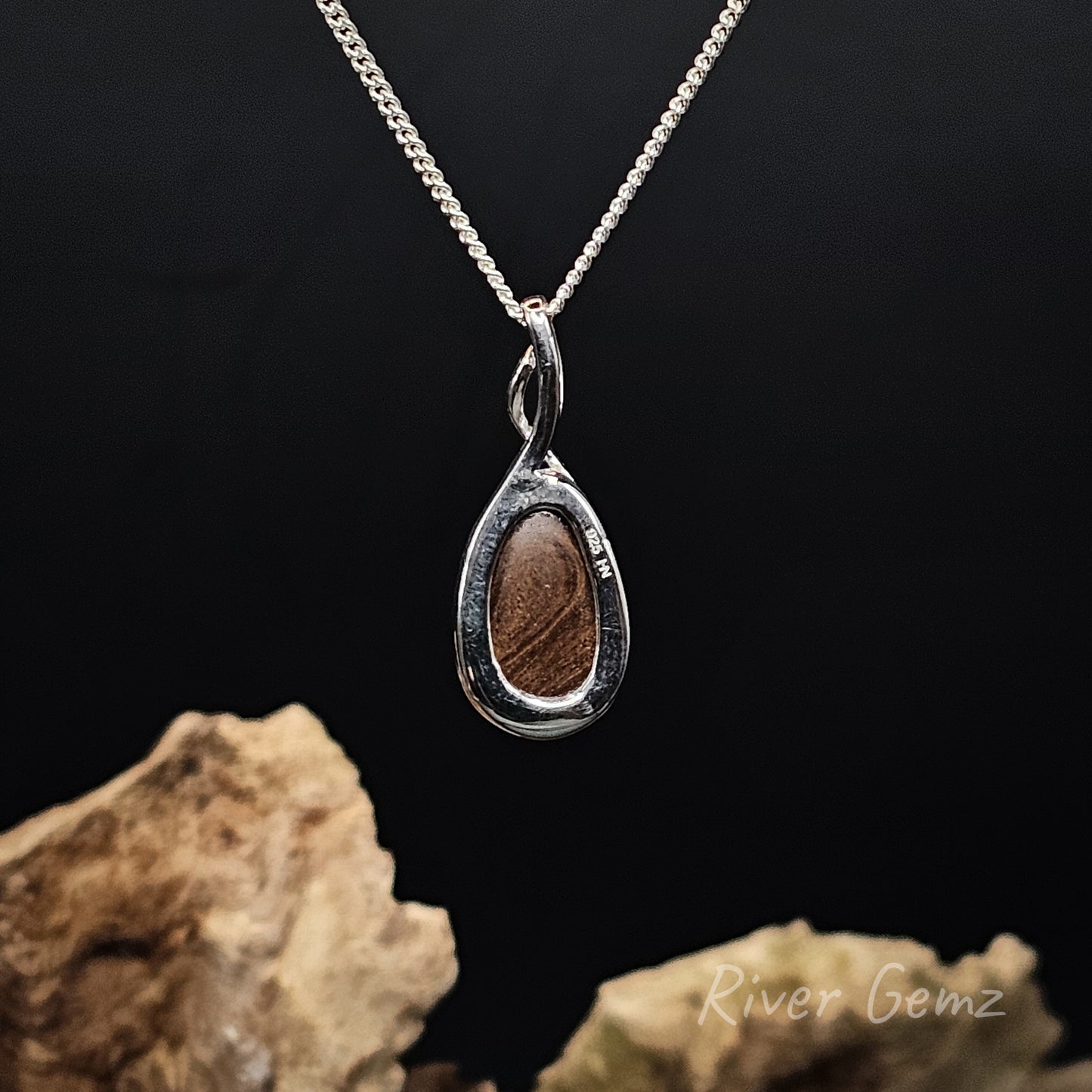 Rear view of a silver teardrop pendant necklace showing the brown ironstone of the included boulder opal, suspended from a silver chain against a dark background with decorative rocks at the bottom.