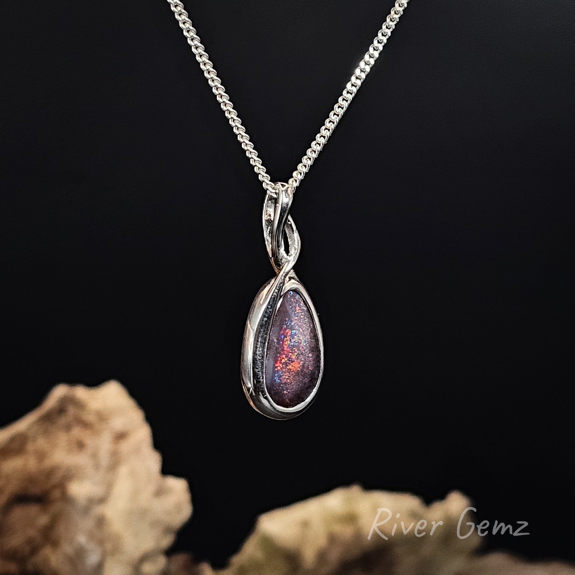 A silver teardrop pendant necklace with a swirling inlay of red, orange, and purple glitter, displayed on a black background with decorative rocks at the bottom.