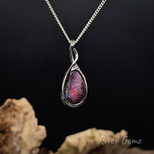 A silver teardrop pendant necklace with a swirling iridescent opal-like gemstone showing pink, purple, and blue flashes, displayed on a black background with decorative rocks at the bottom and a 'River Gemz' watermark.