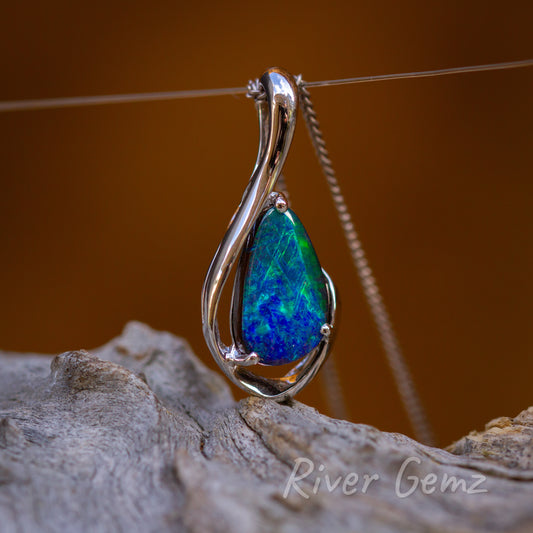 Blue-green solid opal held securely by 3 claws in the white gold pendant. The bail is inconspicuosly part of the pendant design which was based on a fish hook. Pendant sits above a weathered light grey piece of drift wood and a burnt orange backdrop.