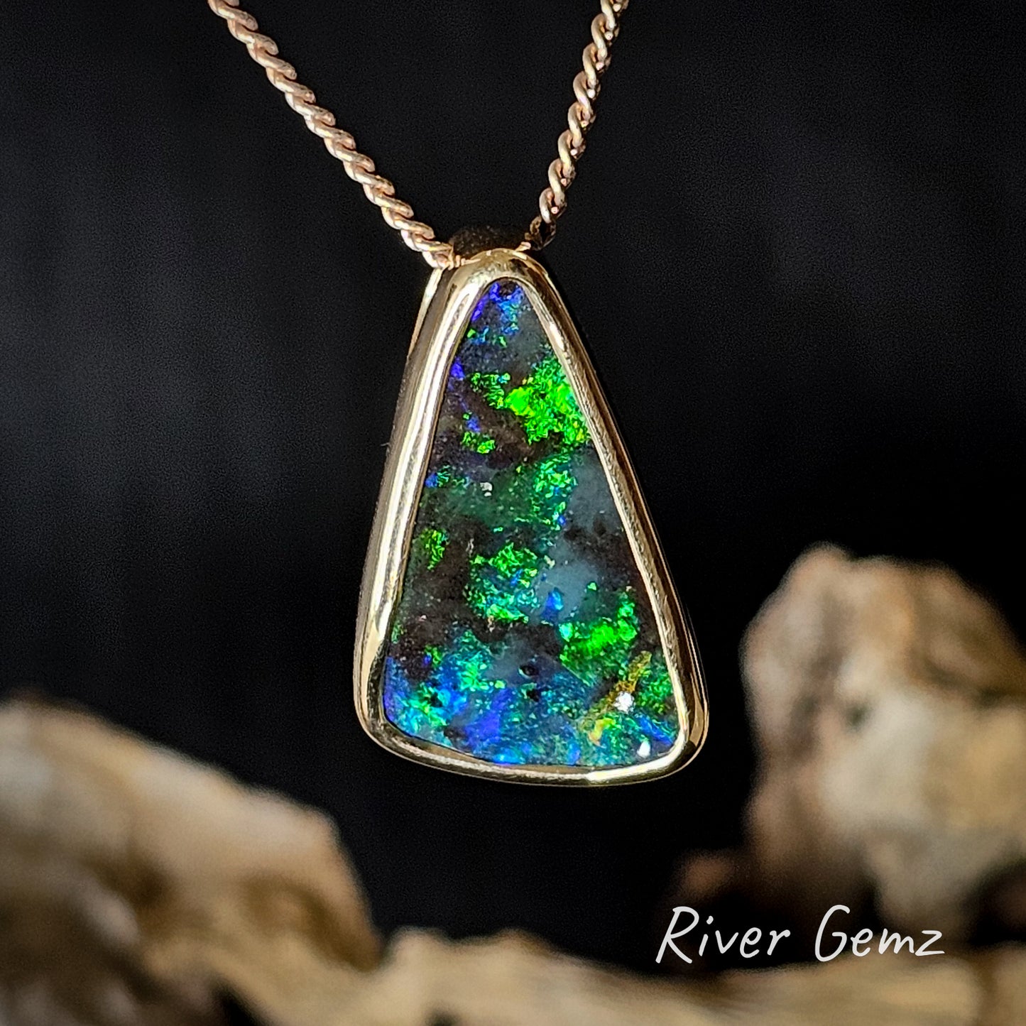 Triangular shaped colourful boulder opal besel set in yellow gold pendant on a dark background, featuring 'River Gemz' branding.