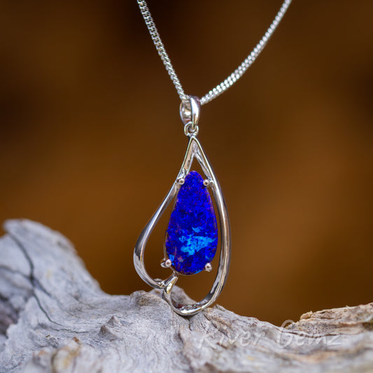 Vivid blue tear shaped opal held securely by 4 claws in this white gold pendant. The setting resembles an inverted love heart with a small bail to complete the design. The image shows the pendant upright sitting above a light-grey grained timber and burnt orange background.