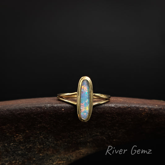 Colourful oval shaped opal besel set in yellow gold ring on a shoe anvil against a dark background and 'River Gemz' watermark in the bottom right hand corner.