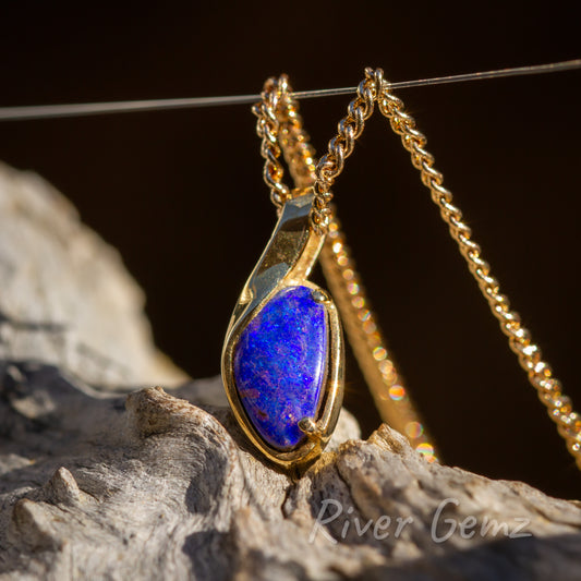 Rounded corners on the triangular shaped opal secured with besel and claw setting in the pendant. Chain passes through a gap created in the pendant design and thus no separate bail. Gold contrasts strongly against the dark brown background and the light grey grain of the driftwood that the pendant sits above.