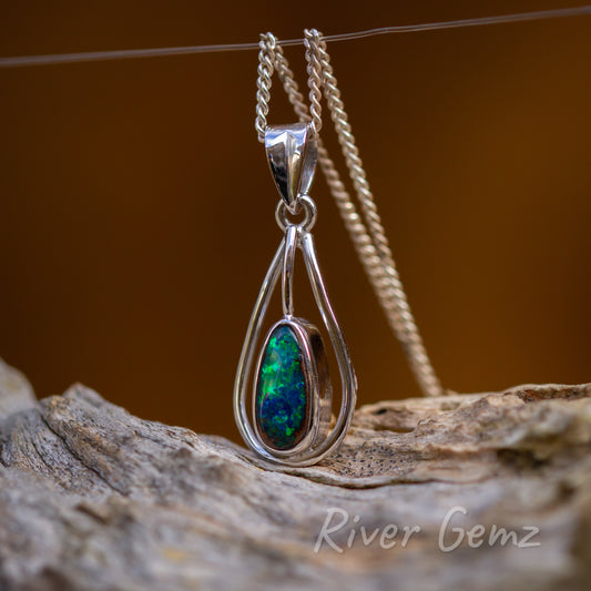 Oval shaped opal is within a tear drop shaped setting with large tapered bail. Pendant is photographed upright with burnt orange back drop and standing on a light grey graine wood piece.