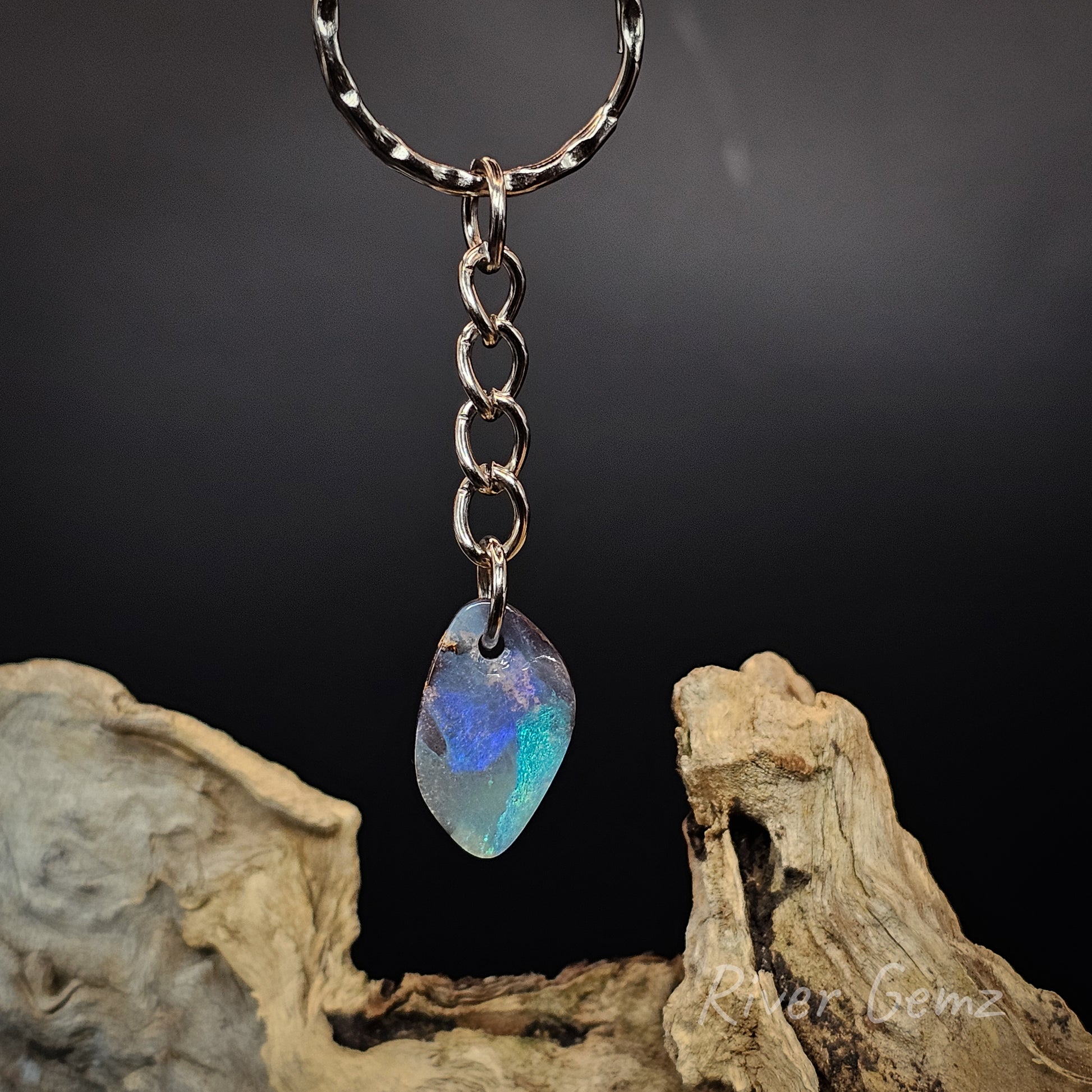 A silver-tone keychain with a teardrop-shaped iridescent blue opal pendant, resting on a piece of driftwood against a dark background.