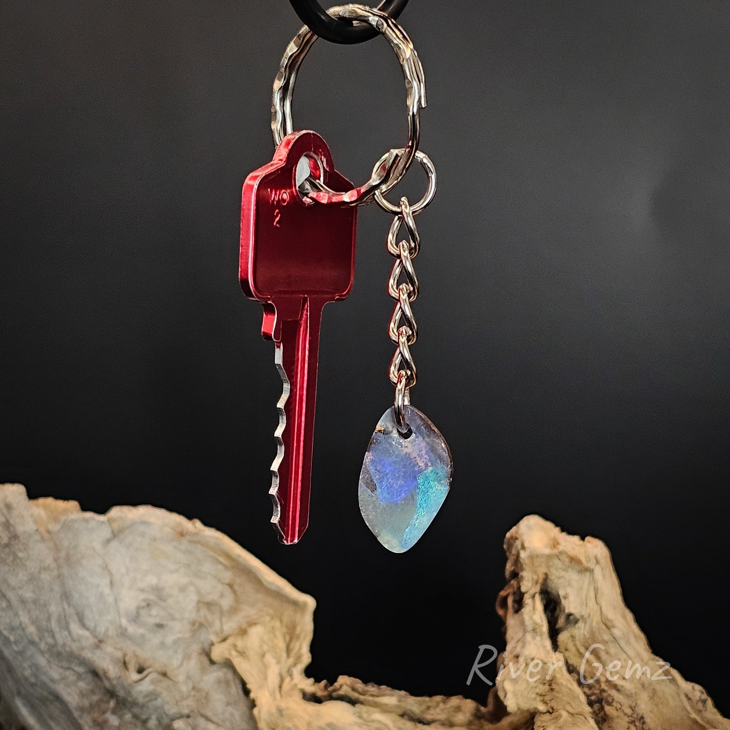 A keychain featuring a red metal key, a silver split ring, and a teardrop-shaped iridescent gemstone charm, displayed against a dark background with driftwood.