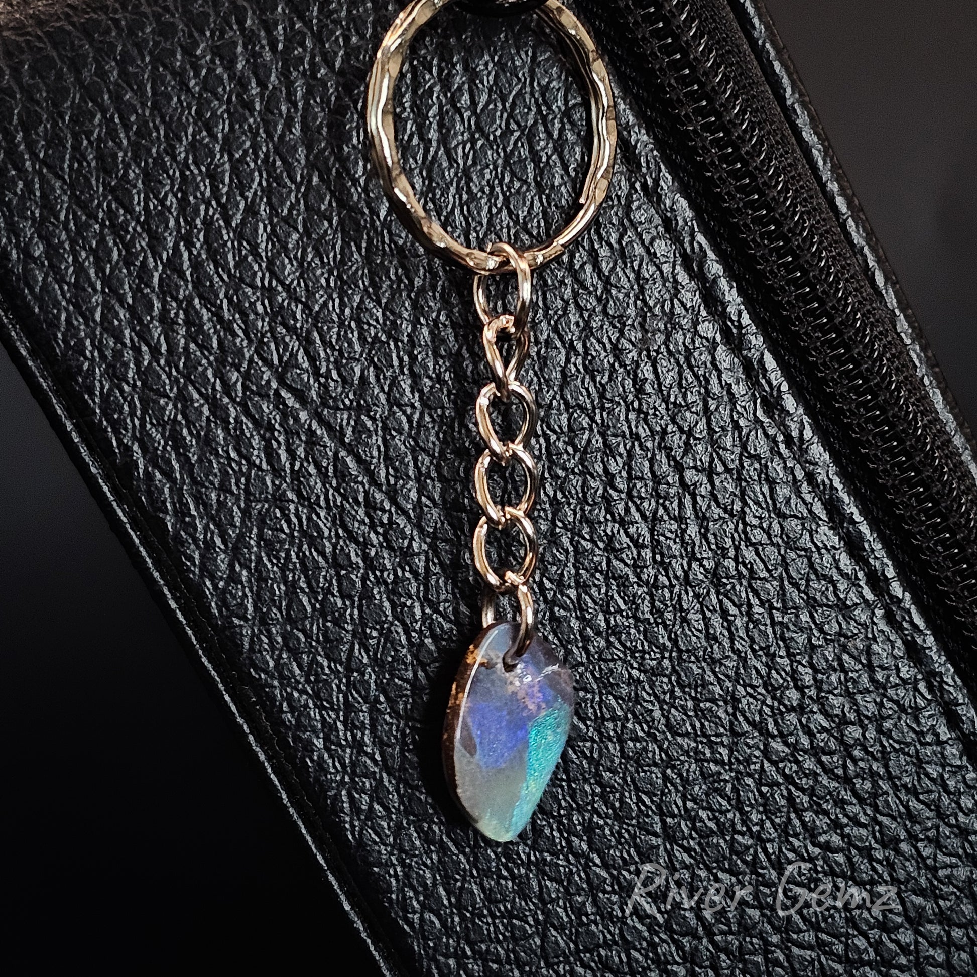 A close-up of a teardrop-shaped iridescent opal bag charm, resting on a black textured leather surface.
