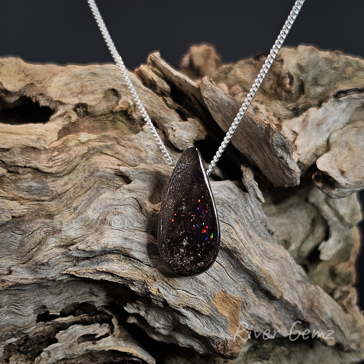 Tear-drop shaped boulder opal with flecks of colour with a silver chain passing through it on a piece of light brown driftwood.