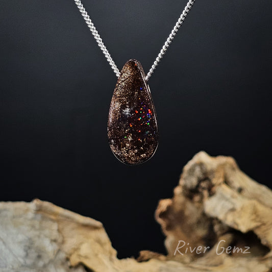 Boulder opal necklace with a silver chain passing through it against a dark background and light driftwood at the base with 'River Gemz' watermark in the bottom right hand corner.