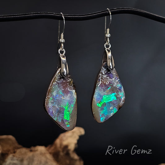 A pair of handmade dangle earrings featuring raw, irregularly shaped opal stones with silver-toned metal findings and fishhook wires, hanging from a black display stand against a dark background. The opal displays vibrant flashes of green, blue, and purple.