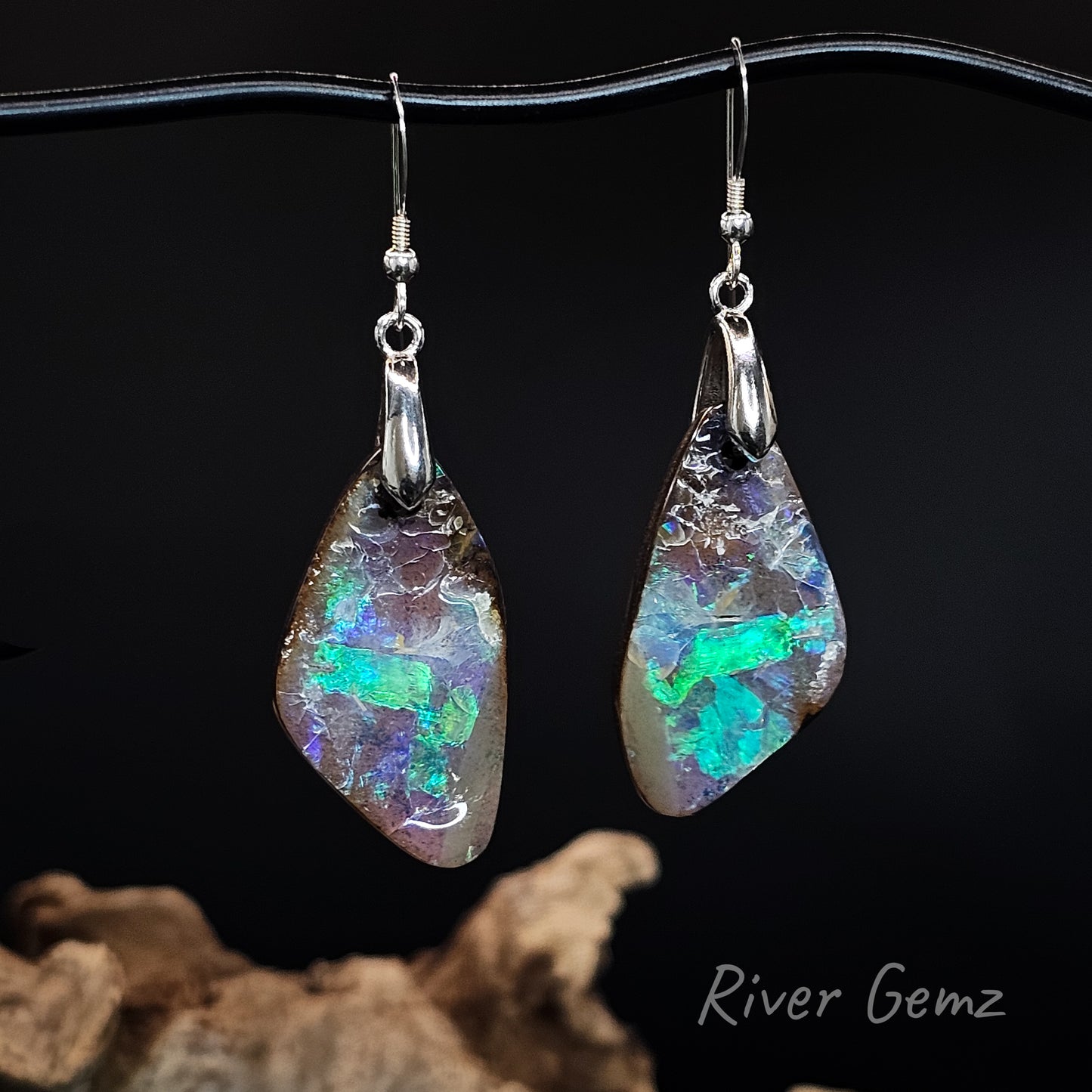 A pair of dangling earrings with irregular teardrop-shaped iridescent opal stones in silver hooks, displayed against a black background with driftwood at the bottom and the text 'River Gemz' in the lower right.