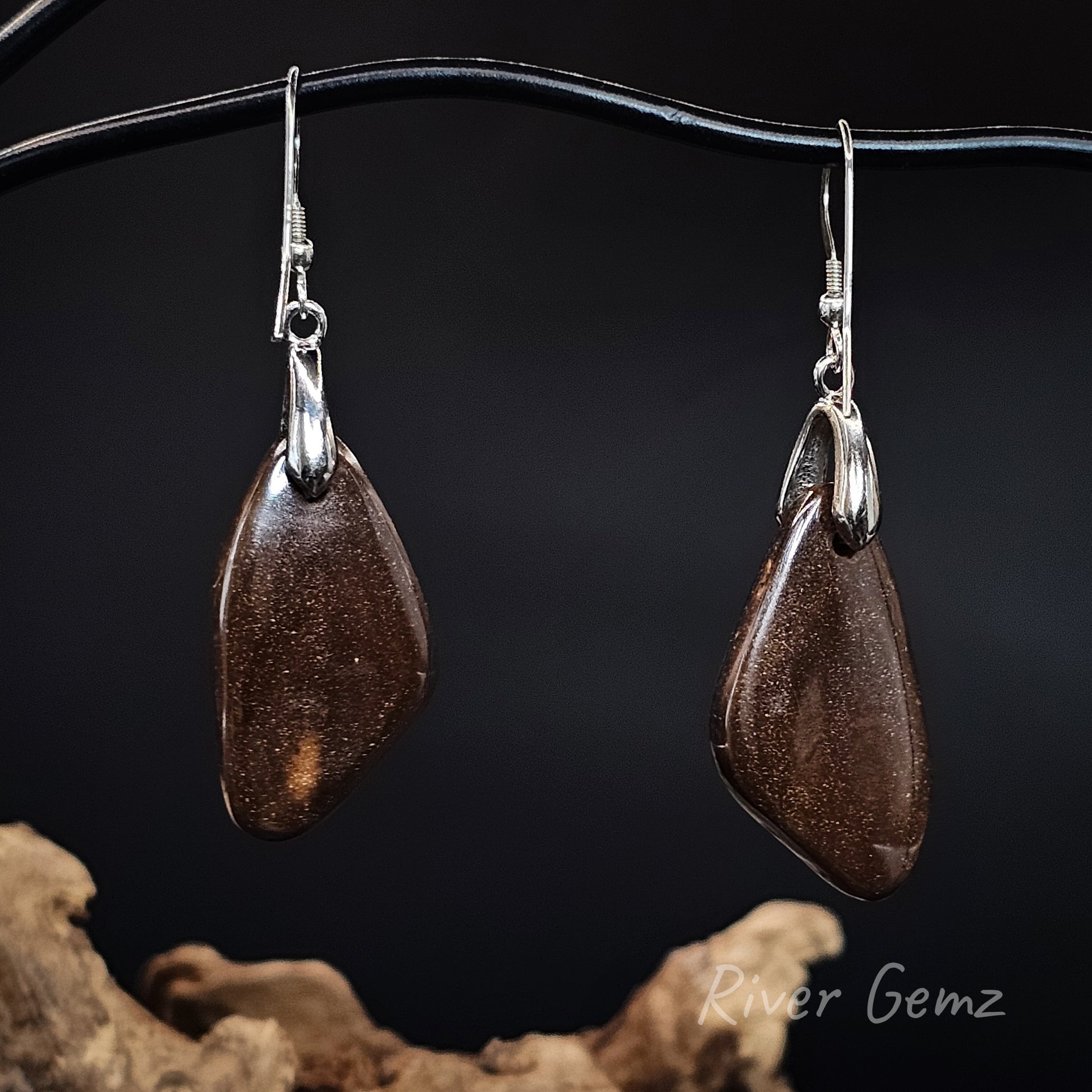 Rear view of a pair of silver drop earrings showing the ironstone matrix of the boulder opals, hanging from a black display stand against a dark background with driftwood at the bottom.